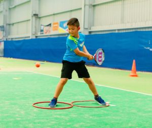 A young person with a foot in separate hoops about to hit a tennis ball