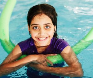 Young person smiling at the camera while holding a noodle in a pool